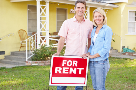 Husband and wife standing in front of house with a for rent sign